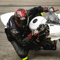A motorcyclist wearing a black leather suit and red-and-black helmet is leaning into a turn while riding a white sport bike. The rider's gloves are black with red accents, and the detailed textures of the suit and bike are visible.