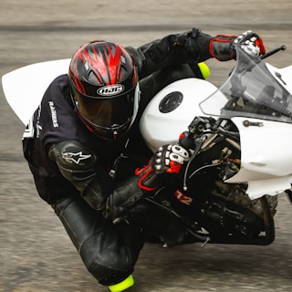 A motorcyclist wearing a black leather suit and red-and-black helmet is leaning into a turn while riding a white sport bike. The rider's gloves are black with red accents, and the detailed textures of the suit and bike are visible.