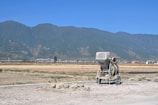 A construction mixer is situated on a partially developed site with rocky terrain in the foreground. In the background, expansive fields stretch out towards a distant town nestled at the base of a range of towering greenish mountains under a clear blue sky.