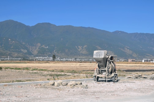 A construction mixer is situated on a partially developed site with rocky terrain in the foreground. In the background, expansive fields stretch out towards a distant town nestled at the base of a range of towering greenish mountains under a clear blue sky.