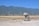 A construction mixer is situated on a partially developed site with rocky terrain in the foreground. In the background, expansive fields stretch out towards a distant town nestled at the base of a range of towering greenish mountains under a clear blue sky.