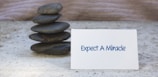A small stack of smooth, dark stones is arranged neatly on a textured surface. Next to the stones is a white card displaying the text 'Expect A Miracle' in blue letters. The background has a subtle wavy pattern, adding to the calm and serene atmosphere of the scene.