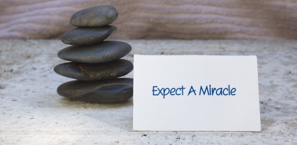 A small stack of smooth, dark stones is arranged neatly on a textured surface. Next to the stones is a white card displaying the text 'Expect A Miracle' in blue letters. The background has a subtle wavy pattern, adding to the calm and serene atmosphere of the scene.