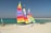 Three brightly colored catamarans are situated on a sandy beach. The sails feature vivid stripes in colors such as yellow, red, orange, purple, and green. The turquoise sea and clear blue sky create a serene backdrop. A jetty of rocks stretches into the sea, and another small sailing vessel is visible in the distance along with a trailer on the beach.