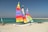 Three brightly colored catamarans are situated on a sandy beach. The sails feature vivid stripes in colors such as yellow, red, orange, purple, and green. The turquoise sea and clear blue sky create a serene backdrop. A jetty of rocks stretches into the sea, and another small sailing vessel is visible in the distance along with a trailer on the beach.