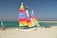 Three brightly colored catamarans are situated on a sandy beach. The sails feature vivid stripes in colors such as yellow, red, orange, purple, and green. The turquoise sea and clear blue sky create a serene backdrop. A jetty of rocks stretches into the sea, and another small sailing vessel is visible in the distance along with a trailer on the beach.