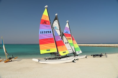 Three brightly colored catamarans are situated on a sandy beach. The sails feature vivid stripes in colors such as yellow, red, orange, purple, and green. The turquoise sea and clear blue sky create a serene backdrop. A jetty of rocks stretches into the sea, and another small sailing vessel is visible in the distance along with a trailer on the beach.