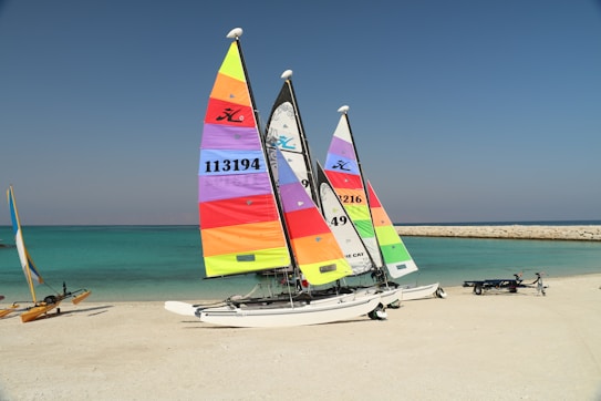 Three brightly colored catamarans are situated on a sandy beach. The sails feature vivid stripes in colors such as yellow, red, orange, purple, and green. The turquoise sea and clear blue sky create a serene backdrop. A jetty of rocks stretches into the sea, and another small sailing vessel is visible in the distance along with a trailer on the beach.