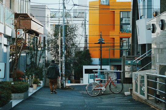 A narrow urban street with modern architecture and a mix of old and new buildings. A person in a hoodie walks down the street, and a bright orange building stands out in the background. A red bicycle is parked against a metal railing near the center, while various plants and urban elements like utility poles and cables add character to the scene.