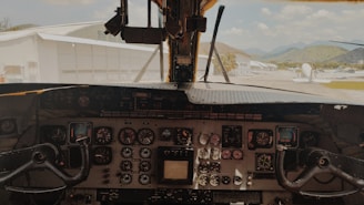 A cockpit view of an aircraft with various instruments and controls visible on the panel. There are analog dials, switches, and two control yokes for piloting the plane. Outside the cockpit window, hangars, an aircraft, and distant hills under a partly cloudy sky can be seen.