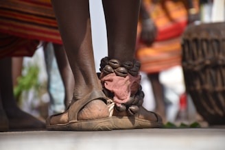 A close-up of a person's leg adorned with traditional ankle jewelry made of stones. The person is wearing sandals, and the background shows colorful garments and what appears to be a large drum.