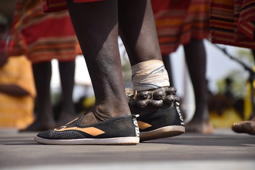 Close-up of a person's lower legs wearing woven anklets adorned with beads, accompanied by vibrant, patterned clothing in the background. The footwear appears to be black with orange patterns, and the setting suggests an outdoor cultural event.