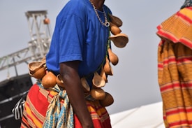 A person is wearing a traditional outfit with a necklace made of small gourds. The attire includes a bright blue shirt and a colorful fabric wrapped around the waist, featuring intricate patterns in red, blue, and yellow. Another person nearby is wearing a garment with orange and red stripes. The background shows a metal structure, possibly part of a stage or event setup.
