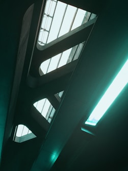 A futuristic interior with a geometric design, featuring large, elongated skylights that allow natural light to enter. The structure consists of sleek, modern lines with a metallic or concrete finish and a greenish tint.