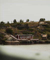 A small wooden house with a dark red facade and white trims is situated by the water's edge. It rests on rocky terrain surrounded by a mix of bushes and small trees. The overcast sky casts a muted light over the scene, contributing to the rustic charm of the isolated countryside setting.