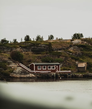 A small wooden house with a dark red facade and white trims is situated by the water's edge. It rests on rocky terrain surrounded by a mix of bushes and small trees. The overcast sky casts a muted light over the scene, contributing to the rustic charm of the isolated countryside setting.