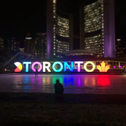 A brightly lit, colorful Toronto sign is displayed in front of a modern building at night. The letters are illuminated in different colors, casting reflections on the ground, with a maple leaf symbol at the end. Silhouettes of people are visible near the sign.