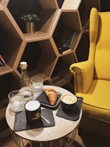 A cozy setting featuring a yellow armchair next to a wooden honeycomb-style bookshelf. On a round table, there is a cup of coffee with foam, a croissant on a slate plate, a glass water bottle, and two glasses. Books and a small potted plant are visible on the shelves.