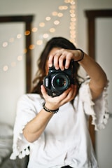 A person dressed in a white shirt is holding a black DSLR camera in front of their face, focusing directly at the camera lens. The background is blurred with a string of warm fairy lights, creating a bokeh effect.