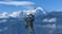 A person is hiking in a mountainous region, wearing outdoor gear and using a camera to photograph the scenery. The background features snow-capped peaks and a clear blue sky.