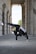 A person is performing a one-handed handstand in a corridor surrounded by large stone columns. The individual is wearing a black athletic outfit with a white stripe and red shoes. The setting gives a sense of urban architecture and outdoor space, with greenery visible in the background.