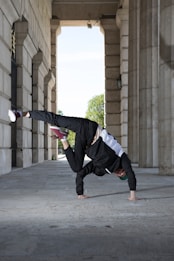 A person is performing a one-handed handstand in a corridor surrounded by large stone columns. The individual is wearing a black athletic outfit with a white stripe and red shoes. The setting gives a sense of urban architecture and outdoor space, with greenery visible in the background.
