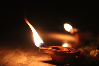 A detailed close-up of an earthen diya lamp with a bright flame, casting a warm glow. The lamp is placed on a reflective surface, and there is another diya visible in the background, slightly out of focus. Some scattered flowers can be seen at the edge, adding to the festive ambiance.