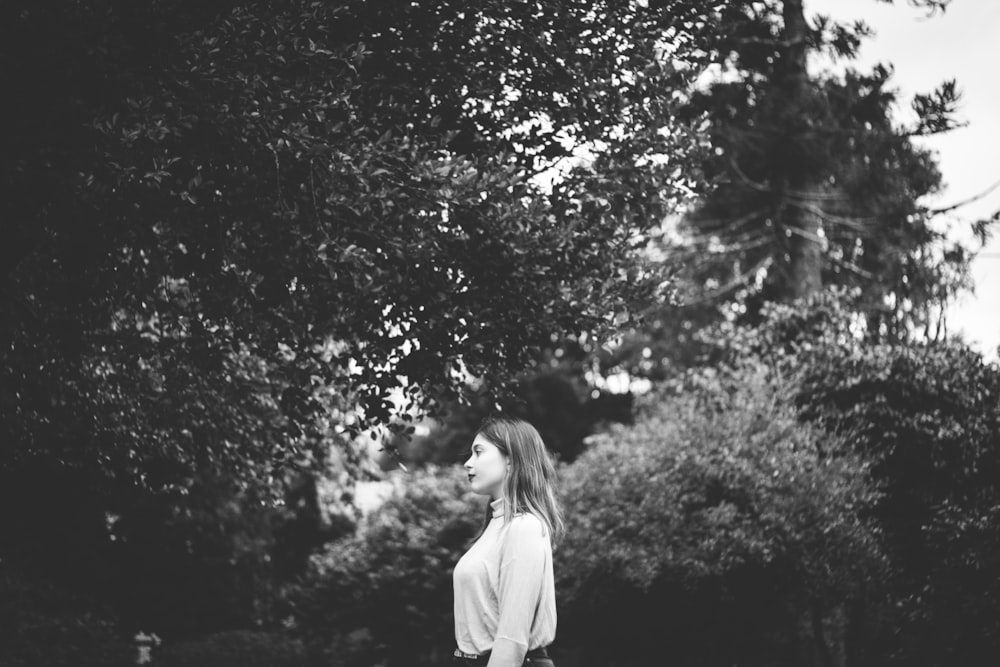 grayscale photography of standing woman in long-sleeved shirt beside tree