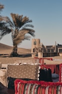 A serene desert setting featuring traditional seating arrangements with decorative cushions, a tall palm tree, and an adobe-style structure in the background. The sandy landscape and clear blue sky create a peaceful atmosphere.