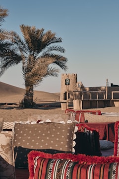 A serene desert setting featuring traditional seating arrangements with decorative cushions, a tall palm tree, and an adobe-style structure in the background. The sandy landscape and clear blue sky create a peaceful atmosphere.