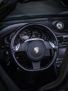 A sleek and modern car interior showcasing a black steering wheel with the Porsche emblem displayed prominently in the center. The dashboard features various gauges and dials, and there are subtle metallic accents.