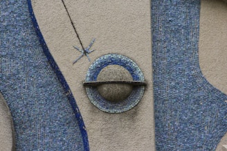 A textured wall mural features abstract designs composed of small blue tiles arranged in flowing patterns. The centerpiece resembles a ring or planet with a single line crossing horizontally. A small star-like design is positioned above the central object.