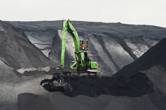 A large green excavator is perched on the edge of a steep, dark, rocky slope, seemingly involved in mining or excavation work. The machine stands out against the gray and black tones of the terrain, with layers of rock visible. A person is visible inside the cabin of the excavator.