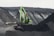 A large green excavator is perched on the edge of a steep, dark, rocky slope, seemingly involved in mining or excavation work. The machine stands out against the gray and black tones of the terrain, with layers of rock visible. A person is visible inside the cabin of the excavator.