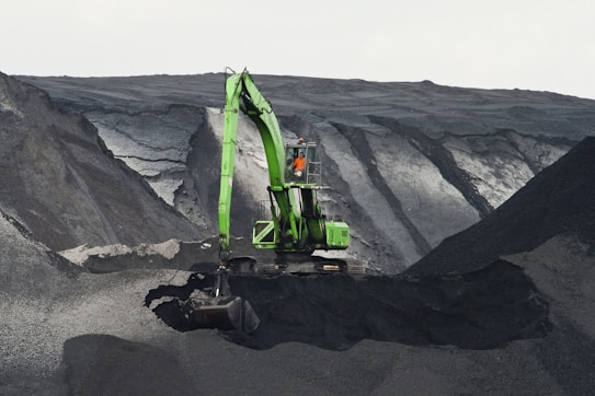 A large green excavator is perched on the edge of a steep, dark, rocky slope, seemingly involved in mining or excavation work. The machine stands out against the gray and black tones of the terrain, with layers of rock visible. A person is visible inside the cabin of the excavator.