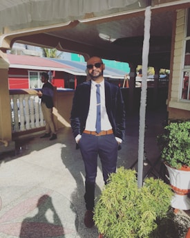 A man in formal attire stands confidently outdoors. He wears a dark blazer, white shirt, and a tie, with hands in his pockets. Sunglasses add a casual touch, while sunlight casts shadows on a textured floor. A second person can be seen in the background near a white railing and red building.