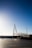 A modern pedestrian bridge with sleek, white support cables spans over a body of water. The bridge features a triangular support structure and a curved walkway. Below, the tiles of a plaza reflect light from the setting sun. In the background, the horizon and sea are visible, with a few docked sailboats on the left.