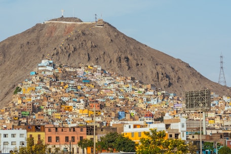 A large, colorful hillside community is densely populated with an array of small houses in various pastel and earthy tones. The dwellings are tightly packed, climbing up the slopes of a barren, rocky hill. A large cross is positioned at the top of the hill, and electrical towers are visible to the side. At the base, some taller buildings and greenery can be seen, suggesting an urban setting.