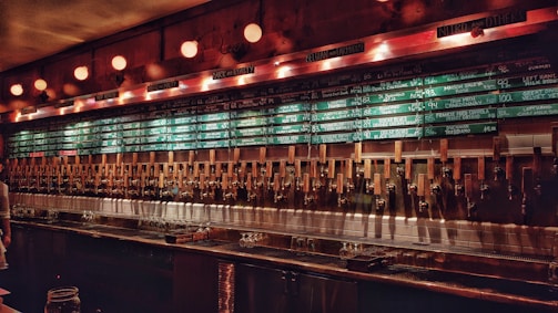 A dimly lit bar with a long wall of taps, showcasing a wide variety of beers. Above the taps, several chalkboards display the available beer selections along with their prices. The ambiance is warm and inviting, with vintage-style light fixtures providing subtle illumination.