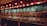 A dimly lit bar with a long wall of taps, showcasing a wide variety of beers. Above the taps, several chalkboards display the available beer selections along with their prices. The ambiance is warm and inviting, with vintage-style light fixtures providing subtle illumination.