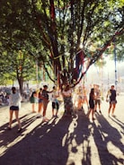 A lively outdoor gathering under a large, leafy tree decorated with colorful ribbons that catch sunlight filtering through the branches. Groups of people, both standing and walking around, are interacting with each other in a festive atmosphere.