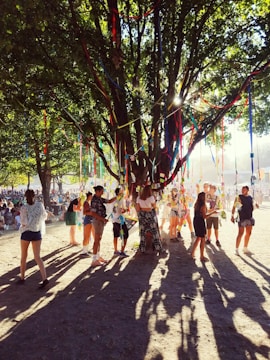 A lively outdoor gathering under a large, leafy tree decorated with colorful ribbons that catch sunlight filtering through the branches. Groups of people, both standing and walking around, are interacting with each other in a festive atmosphere.