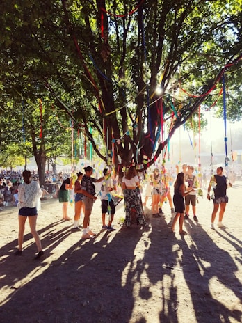 A lively outdoor gathering under a large, leafy tree decorated with colorful ribbons that catch sunlight filtering through the branches. Groups of people, both standing and walking around, are interacting with each other in a festive atmosphere.
