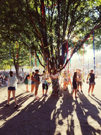 A lively outdoor gathering under a large, leafy tree decorated with colorful ribbons that catch sunlight filtering through the branches. Groups of people, both standing and walking around, are interacting with each other in a festive atmosphere.