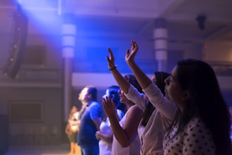 Several people are standing in a dimly lit interior space with soft blue and purple lighting. They appear to be engaged in an activity involving raised hands, possibly singing or worshipping. The background features architectural elements like columns and railings, indicating a large hall or auditorium.