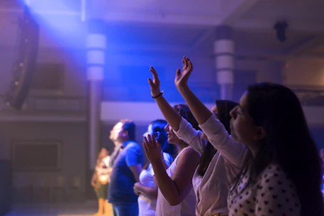A vibrant worship session with hands raised, bathed in soft purple and blue stage lighting.