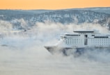 A large cruise ship sails through a misty, wintery landscape with snow-covered hills and trees. The soft orange hue of the sky suggests early morning or late afternoon light.