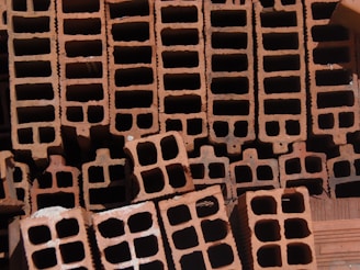 A close-up view of a stack of orange-brown rectangular hollow bricks, arranged in a pattern. The bricks have multiple square cavities running through them, designed for construction purposes. The lighting casts shadows within the cavities, highlighting the texture of the brick surface.