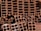 A close-up view of a stack of orange-brown rectangular hollow bricks, arranged in a pattern. The bricks have multiple square cavities running through them, designed for construction purposes. The lighting casts shadows within the cavities, highlighting the texture of the brick surface.