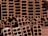 A close-up view of a stack of orange-brown rectangular hollow bricks, arranged in a pattern. The bricks have multiple square cavities running through them, designed for construction purposes. The lighting casts shadows within the cavities, highlighting the texture of the brick surface.